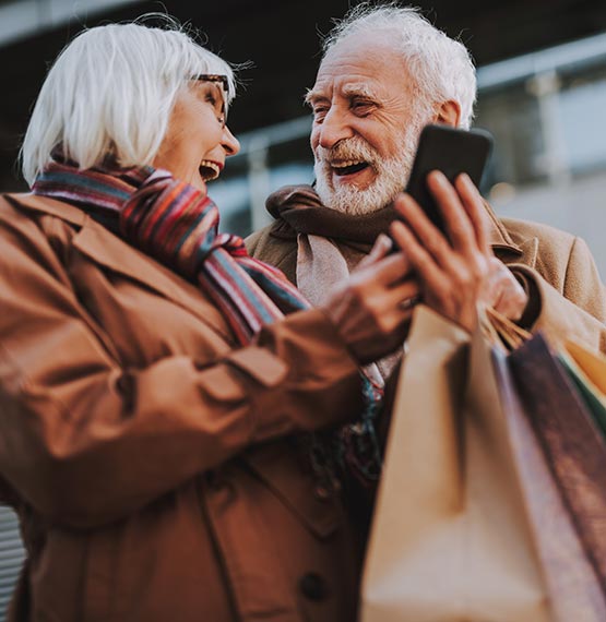 elderly couple with mobile phone smiling at each other while walking with shopping bags in hand