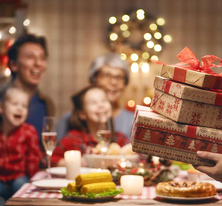 person holding Christmas presents in front of blurred image of happy family sitting for Christmas dinner at table