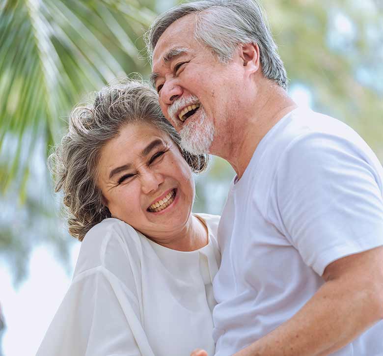 retired Asian man and woman smiling while embracing with blurred palm leaves in the background on what appears to be a tropical beach