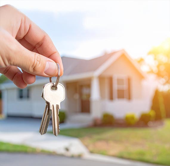 A hand is holding house keys in the foreground. In the background, softly blurred, is a small, manufactured or modular home during golden hour.